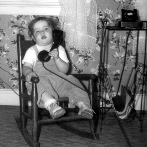 Old picture of baby Pam talking on a rotary telephone. She is sitting in a rocking chair.